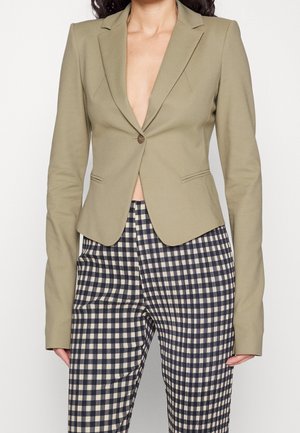 Vrouw draagt een nauwsluitend kaki colbert met een enkele knoop, gecombineerd met zwart-wit geruite, hooggetailleerde broek.