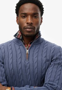 Niebieski sweter w splot warkoczowy z zamkiem na kołnierzu, który ma wytłoczaną podszewkę w kratkę. Sweter ma ściągacze na rękawach oraz subtelną detalizację z logo.