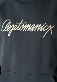 Sudadera azul marino con un gran texto bordado en color crema que dice "Cleptomanicx" en el pecho. El material parece suave y tiene una textura lisa.