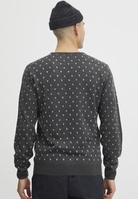 Pull en tricot gris avec des motifs géométriques blancs, col rond, manches longues, et bords côtelés au niveau des poignets et de l'ourlet. Porté avec un bonnet noir.