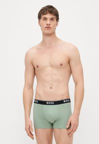 Cuecas boxer verdes de algodão com uma faixa preta na cintura exibindo o logotipo "BOSS". Modelo posa em pé contra um fundo branco simples.