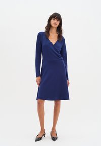 Navy blue wrap dress with long sleeves, V-neckline, and a flared skirt. Made of smooth fabric with a soft texture. Pair of black heeled shoes.