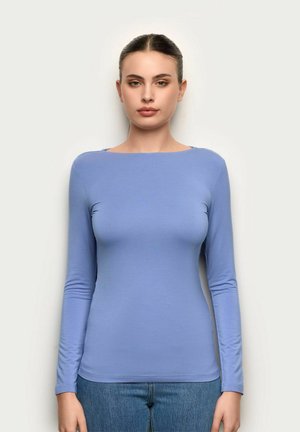 Kobieta o ciemnych włosach związanych do tyłu, ubrana w dopasowaną, jasnoniebieską bluzkę z długim rękawem i niebieskie dżinsy, stojąca na tle jednolitego, jasnego tła.