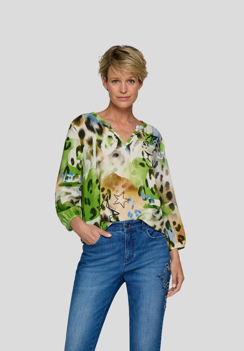 Frau mit einer bunten blauen und grünen Bluse mit Sternen und dem Wort "Love", kombiniert mit blauen Jeans, die Sternapplikationen aufweisen.