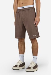 Shorts marron côtelés avec une taille élastique et des poches latérales, arborant un logo "Pegador" blanc. Portés avec des chaussettes blanches à la cheville et des baskets.
