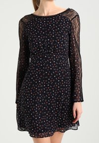 Langärmeliges schwarzes Kleid mit Blumenmustern in Rot, Blau und Weiß. Transparente Spitzenärmel, taillierter Bund und fließender Rock.