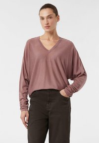 T-shirt z dekoltem w serek, długim rękawem, wykonany z miękkiego materiału w kolorze mauve; luźny krój z marszczonymi mankietami, w połączeniu z ciemno-brązowymi spodniami; minimalistyczny design.