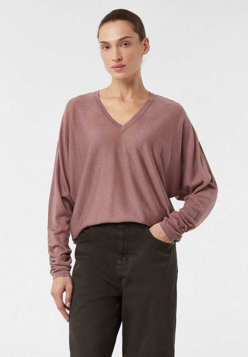 T-shirt z dekoltem w serek, długim rękawem, wykonany z miękkiego materiału w kolorze mauve; luźny krój z marszczonymi mankietami, w połączeniu z ciemno-brązowymi spodniami; minimalistyczny design.