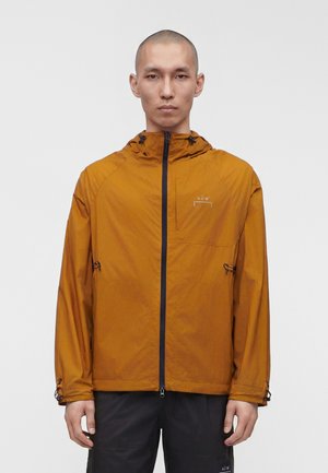 Mand iført en sennepsgul lynlåstrøje med hætte og sorte bukser mod en ensfarvet hvid baggrund, stående stille og vendt fremad.