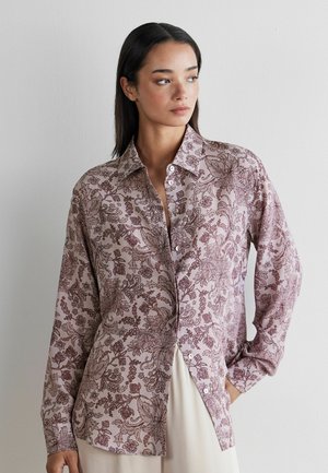 Kobieta ubrana w długą, fioletową koszulę na guziki z ciemnymi kwiatowymi wzorami, patrząca na bok z jedną ręką w kieszeni.