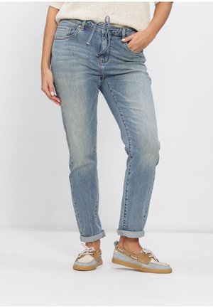 Femme portant un jean bleu clair retroussé avec une taille à cordon et des chaussures bateau beige et grises, debout avec une main dans la poche.