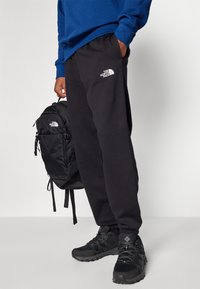 Sort joggingbukser med hvidt logo, blå crewneck sweatshirt, sort rygsæk og sorte træningssko med teksturerede såler.