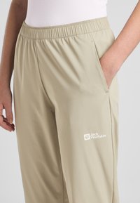Beige lichtgewicht broek met een elastische tailleband en zijzak, met een wit Jack Wolfskin-logo op het linkerbeen. Glad van structuur.