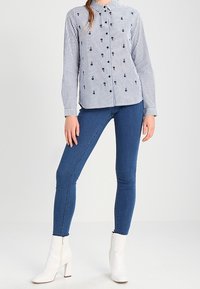 Chemise à boutons rayée avec lignes bleu marine et blanches, détails brodés, associée à un jean skinny bleu et à des bottines blanches à fermeture éclair.