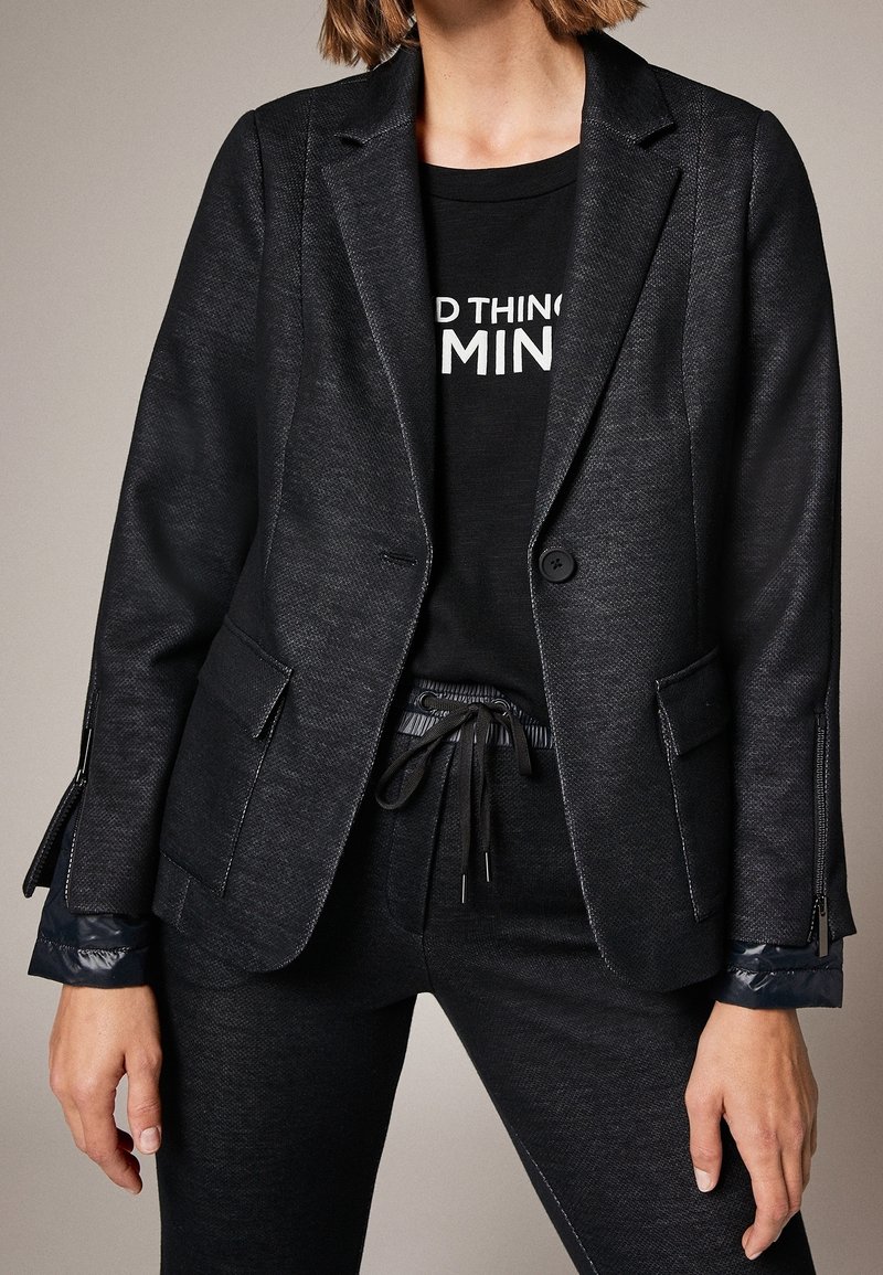 Femme portant un blazer noir ajusté texturé avec poches à rabat, poignets zippés et pantalon noir à cordon sur un t-shirt graphique partiellement visible.
