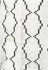 Άσπρο ύφασμα με υφή ανοιχτού πλεκτού, που διαθέτει κάθετα μαύρα μοτίβα σε σχήμα ρόμβου και μικρά άσπρα κουμπιά στη μία πλευρά.