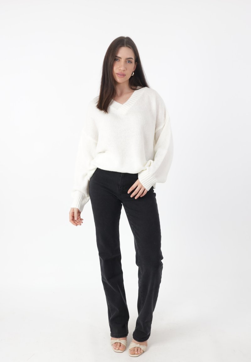 Witte, oversized v-hals trui met geribde manchetten, gecombineerd met zwarte jeans en beige haksandalen, staand voor een effen achtergrond.