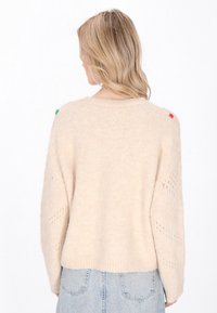 Maglione color crema con vestibilità ampia, lavorazione a maglia testurizzata e motivo a rombi sulle maniche. Presenta piccoli pompon colorati sulle spalle.