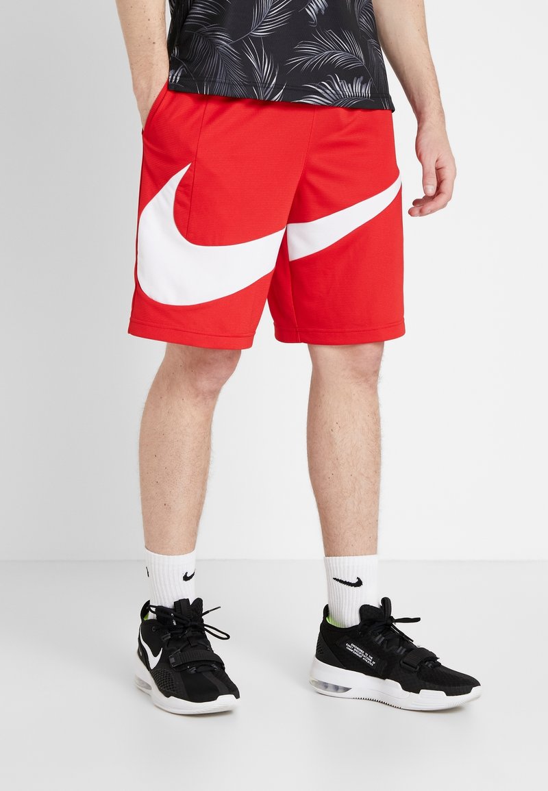 Homme portant un short rouge avec un grand logo Nike blanc, des chaussures de sport noires et blanches, des chaussettes Nike blanches et une chemise noire à motif de feuilles.