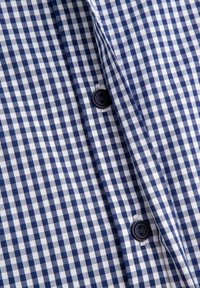 Koszula w kratę w kolorach granatowym i białym z krótkimi rękawami, zapinana na guziki, o gładkiej fakturze tkaniny. Widoczny szczegół guzika i kołnierzyk.