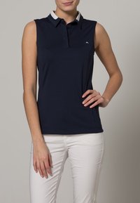 Laivastonsininen hihaton poolopaita, jossa on valkoinen kaulus, nappilista ja pieni logo vasemmassa rinnassa. Tasainen, joustava kangas.