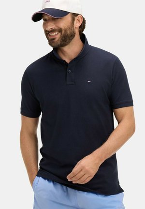 Homme souriant, portant un polo bleu marine, un short bleu clair et une casquette de baseball blanche avec une visière bleu marine, debout devant un fond clair uni.