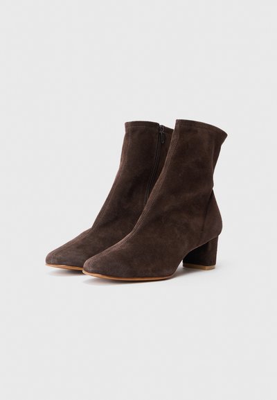 Un paio di stivaletti alla caviglia in suede marrone scuro con zip laterali e tacco basso, su uno sfondo bianco.