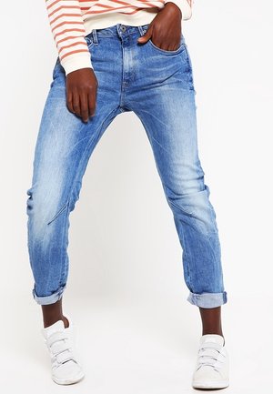 Persoon met opgerolde lichtblauwe spijkerbroek, witte sneakers en een crème-wit met rode gestreepte lange mouwen shirt, staand met één hand in de zak.