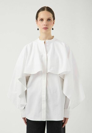 Kvinde med trukket hår tilbage bærer en stor hvid bluse med brede, kapelignende ærmer og sorte hængeøreringe mod en ensfarvet baggrund.