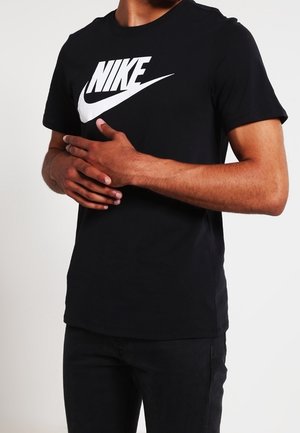 Mężczyzna w czarnej koszulce Nike z białym logo, dłonie złączone na talii, stojący na jednolitym jasnym tle.