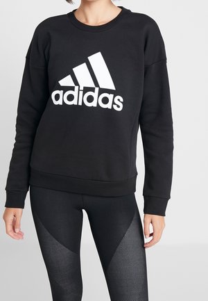 Personne portant un sweat-shirt noir Adidas avec un grand logo blanc et un legging noir texturé sur un fond uni.
