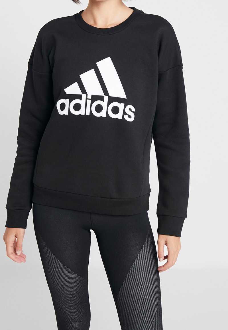 Personne portant un sweat-shirt noir Adidas avec un grand logo blanc et un legging noir texturé sur un fond uni.