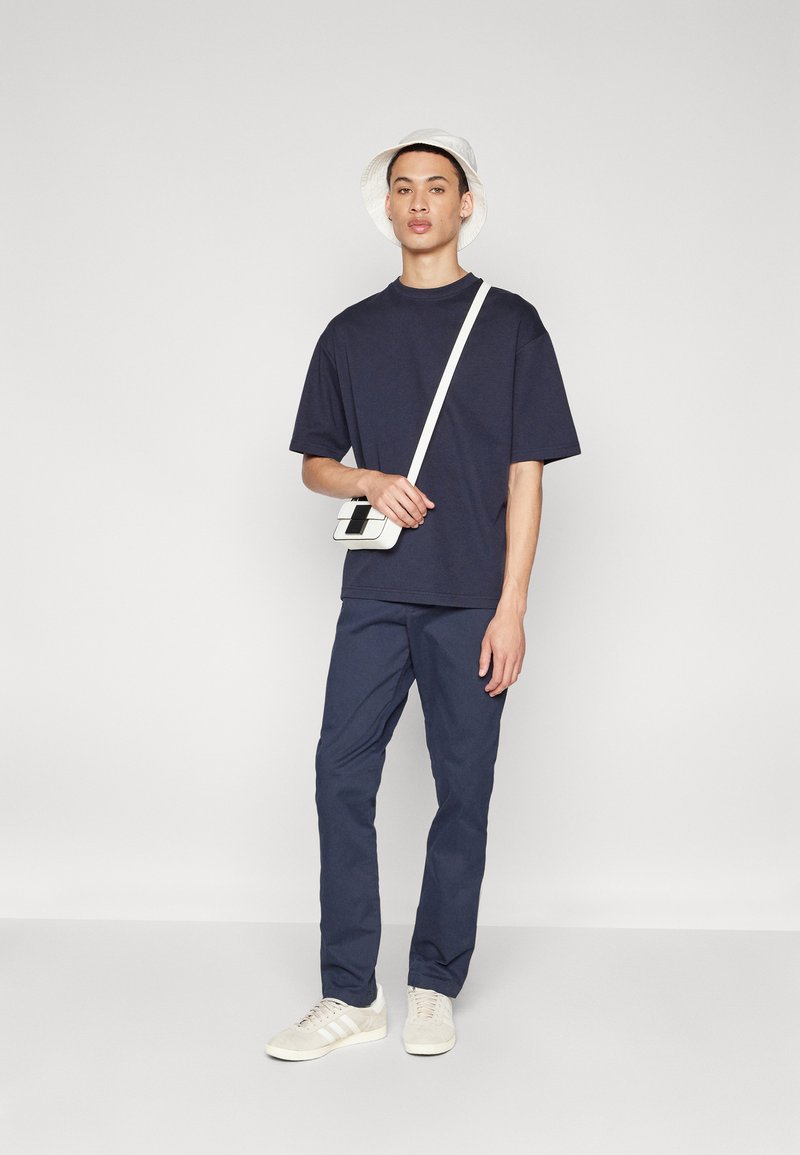 T-shirt oversize bleu marine, pantalon assorti, baskets de couleur claire et chapeau seau blanc. Un sac bandoulière blanc est porté.