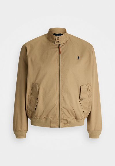 Lett og tynn jakke i beige med stående krage, frontglidelås, elastiske mansjetter og logo på venstre bryst. Har sidelommer og en glatt tekstur.