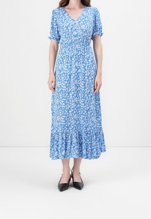 Frau trägt ein blaues V-Ausschnitt-Midikleid mit weißem Blumenmuster, Puffärmeln, geraffter Taille, Stufenrock und schwarzen spitzen Pumps.