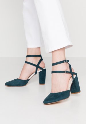Zielone zamszowe buty na wysokim obcasie z wąskim noskiem, z paskami na kostkę i szerokim słupkowym obcasem. Zawierają srebrny akcent na obcasie.