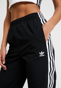 Svarta Adidas träningsbyxor med vita sidostråk, elastisk midja och en liten logotyp på vänster sida. Tillverkade av lättviktig material.