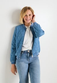 Chaqueta bomber acolchada azul con cierre de cremallera, puños acanalados y cuello redondo, llevada sobre una camisa blanca y combinada con jeans claros.