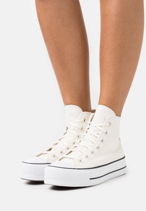 Benen met witte hoge canvas sneakers met dikke witte zolen en zwarte rand, afgebeeld tegen een effen witte achtergrond.