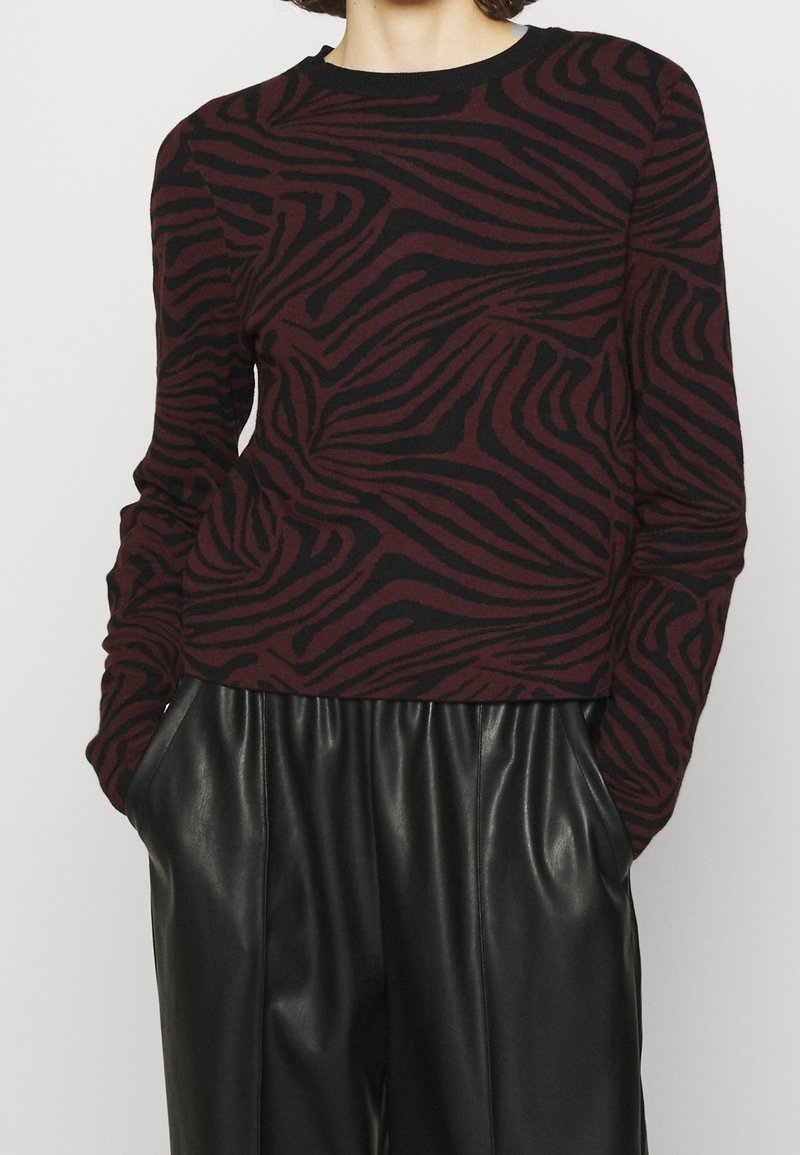 Vínový svetr s černými zebřími pruhy, kulatým výstřihem a dlouhými rukávy, zkombinovaný s černými koženými kalhotami, které mají boční kapsy.