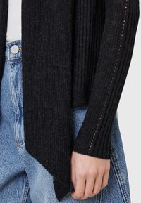 Black knitted cardigan with ribbed texture, featuring delicate cutouts on the sleeves. Paired with light blue denim.