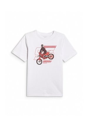 Chłopiec stojący w białej koszulce z czerwonym nadrukiem motocykla, szarym dresie oraz szarych trampkach z różowymi sznurówkami na jednolitym tle.