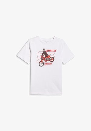 Chłopiec stojący w białej koszulce z czerwonym nadrukiem motocykla, szarym dresie oraz szarych trampkach z różowymi sznurówkami na jednolitym tle.