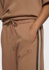 Pantalon de survêtement marron avec taille à cordon, poches latérales, rayures blanches et marron sur le côté, et détail brodé "10".