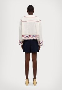 Chaqueta blanca bordada con patrones florales rojos y azules, diseño corto y mangas largas, combinada con pantalones cortos navy y sandalias.