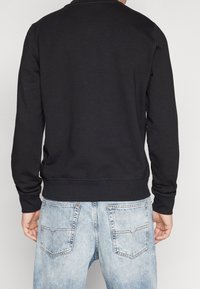 Sudadera negra con puños y dobladillo acanalados, que presenta un escote redondo. Combinada con jeans de mezclilla azul claro, que muestran un diseño distintivo en los bolsillos traseros.