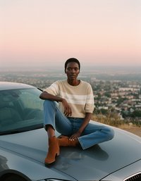 Suéter de punto beige con rayas blancas, jeans azules y botas de tobillo marrones, sentado sobre el capó de un coche plateado con un paisaje urbano desenfocado de fondo.