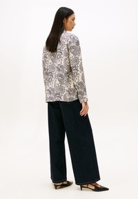 Vrouw staat met een witte paisley-geprinte blouse met lange mouwen, brede donkere jeans en zwarte lage, puntige schoenen, met haar zijwaarts gekeerd.