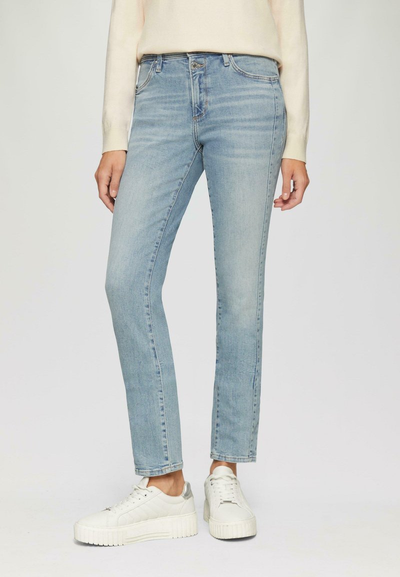 Hellblaue Denim-Jeans mit schlanker Passform, subtilen Verwaschungen, fünf Taschen und einem Reißverschluss. Dazu kombiniert sind weiße Sneakers mit silbernen Akzenten.