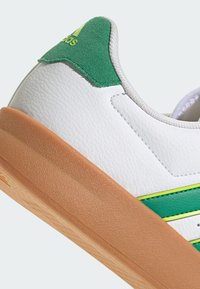 Bijela tenisica s zelenim akcentima, gornjište od teksturirane kože, podstava od smeđe pjene, s žutim prugama i suedenim zelenim tabanom na peti.